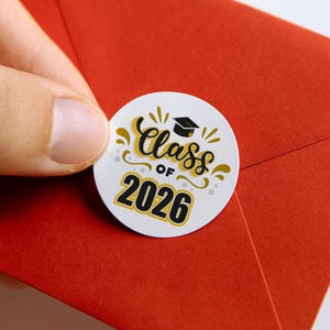 Può includere: Un adesivo rotondo bianco con la scritta "Class of 2026" in nero e oro, con un cappello di laurea. L'adesivo è tenuto da una mano su uno sfondo rosso.