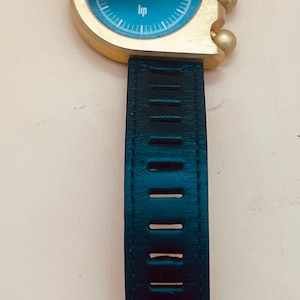 Op de afbeelding: Een polshorloge met een asymmetrische goudkleurige kast en een donkerblauwe wijzerplaat met het Lip-logo. Het horloge heeft drie goudkleurige bolvormige knoppen aan de rechterkant en een donkerblauwe leren band met rechthoekige uitsparingen en een gouden gesp.