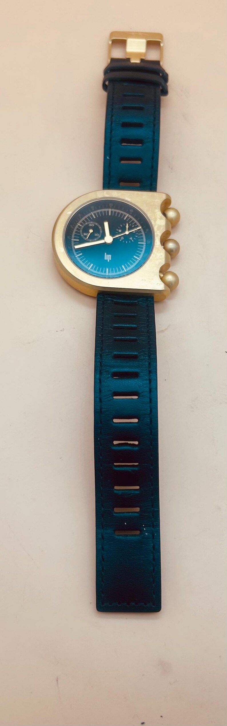 Op de afbeelding: Een polshorloge met een asymmetrische goudkleurige kast en een donkerblauwe wijzerplaat met het Lip-logo. Het horloge heeft drie goudkleurige bolvormige knoppen aan de rechterkant en een donkerblauwe leren band met rechthoekige uitsparingen en een gouden gesp.