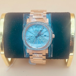 May include: A rose gold wristwatch with a light blue face and a sparkling crystal-studded bezel. The watch has a matching rose gold band and is displayed on a black velvet surface.