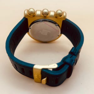 Op de afbeelding: Een polshorloge met een donkerblauwe rubberen band, een goudkleurige gesp en een goudkleurige achterkant van de kast. De horlogekast heeft drie opvallende goudkleurige bolvormige knoppen aan de bovenrand en een ronde metalen achterplaat.