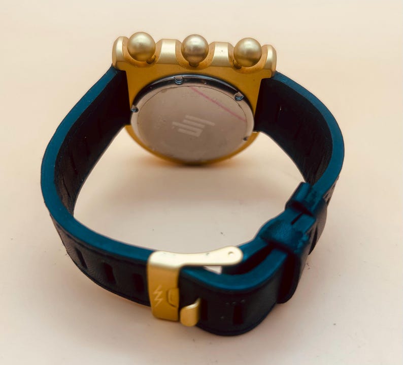 Op de afbeelding: Een polshorloge met een donkerblauwe rubberen band, een goudkleurige gesp en een goudkleurige achterkant van de kast. De horlogekast heeft drie opvallende goudkleurige bolvormige knoppen aan de bovenrand en een ronde metalen achterplaat.