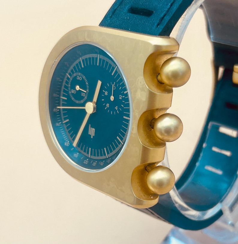Op de afbeelding: Een polshorloge met een ronde blauwgroene wijzerplaat, goudkleurige wijzers en indexen, twee sub-wijzerplaten en het Lip-logo. Het horloge heeft een goudkleurige metalen kast met drie opvallende bolvormige knoppen en een blauwgroene siliconen band.