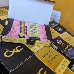 May include: A stack of boxes containing pink embroidery floss. The boxes are black with gold lettering and the text "MADE IN CHINA" on the front. The boxes are labeled with the brand name "CXC" and have a quality assurance seal from SGS.