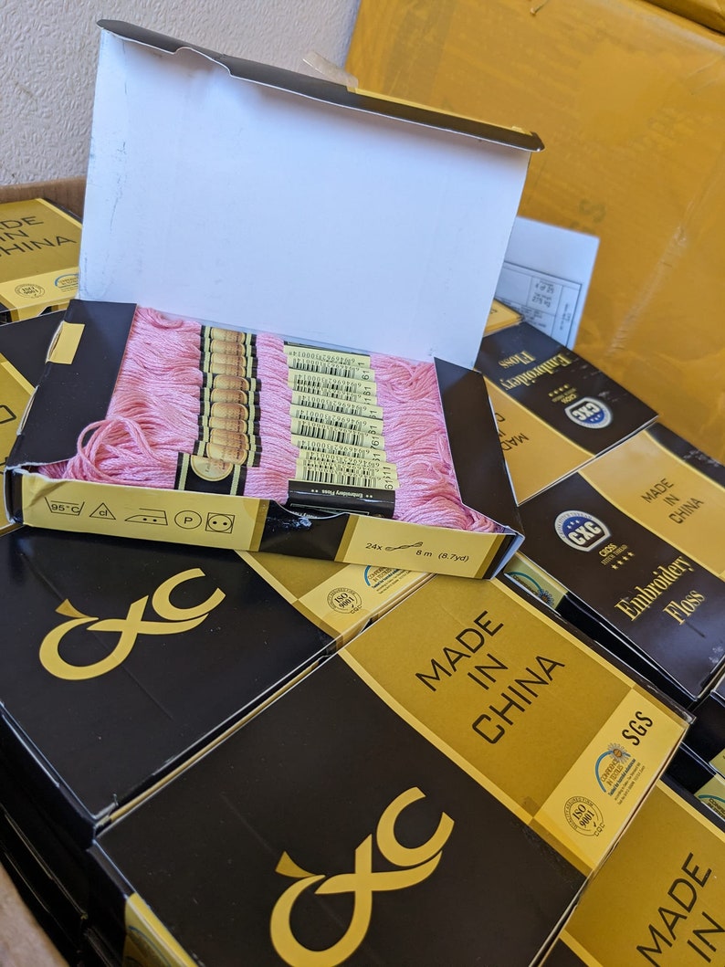 May include: A stack of boxes containing pink embroidery floss. The boxes are black with gold lettering and the text "MADE IN CHINA" on the front. The boxes are labeled with the brand name "CXC" and have a quality assurance seal from SGS.