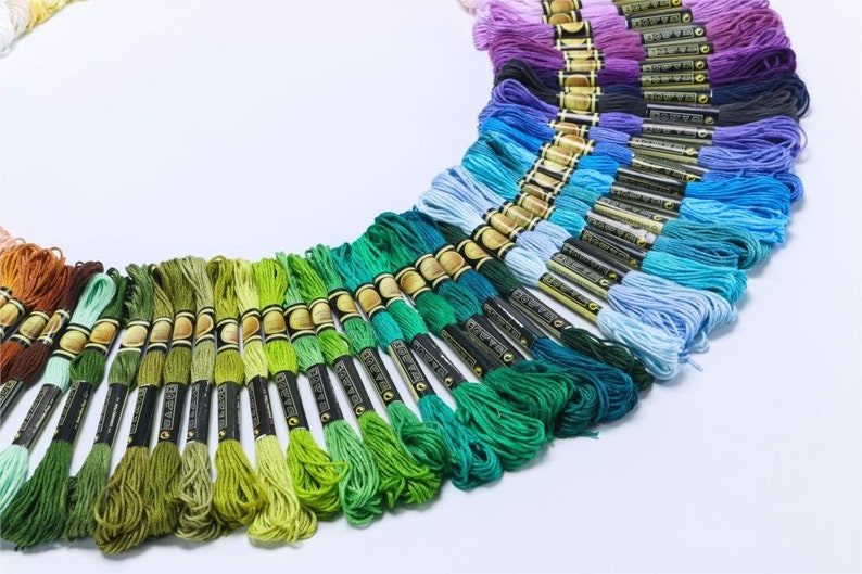 May include: A collection of embroidery floss spools in various shades of green, blue, and purple. The spools are arranged in a semi-circle, with the green shades on the bottom and the purple shades on the top.