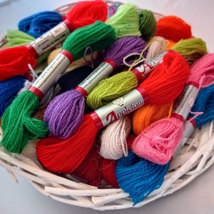 May include: A white woven basket overflowing with colorful embroidery floss. The spools of thread are in shades of red, blue, green, purple, orange, and pink. Each spool has a label that reads "Appleton's" and "100% pure English wool."