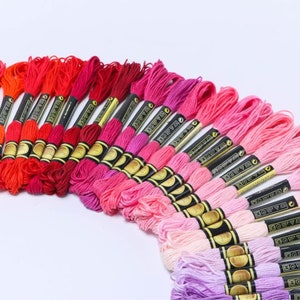 May include: A collection of embroidery floss spools in shades of pink, red, and orange. The floss is arranged in a rainbow gradient, with the darkest shades at the top and the lightest shades at the bottom. Each spool has a black label with gold lettering that reads "DMC" and a number.