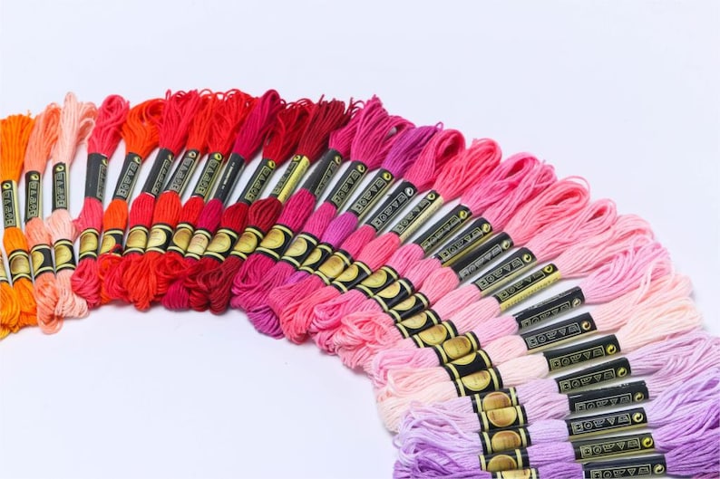 May include: A collection of embroidery floss spools in shades of pink, red, and orange. The floss is arranged in a rainbow gradient, with the darkest shades at the top and the lightest shades at the bottom. Each spool has a black label with gold lettering that reads "DMC" and a number.