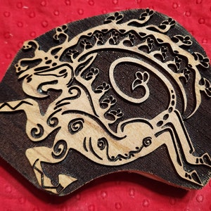 May include: A wooden block carving of a stylized mythical creature with intricate details. The creature has a long, curved tail and a head with horns. The carving is dark brown with light brown details.