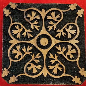 May include: A black wooden block with a carved design of a symmetrical pattern of stylized leaves and a circle in the center. The design is in a light brown color.
