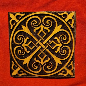 May include: A wooden square tile with a carved design in a dark brown color. The design is a symmetrical pattern of four stylized hearts with intricate details.