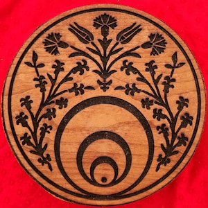 May include: A round wooden coaster with a black floral design and concentric circles in the center. The coaster has a natural wood grain finish.