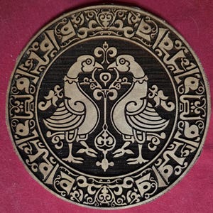 May include: A circular wooden coaster featuring a laser-engraved design of two facing birds in the center. The birds are surrounded by an intricate border of repeating geometric patterns and scrollwork, all rendered in a dark brown and light tan color.