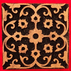 May include: Square wooden block with a carved floral design. The design features a central flower with radiating petals, surrounded by smaller flowers and swirling vines. The wood has a light brown color, contrasting with the black background. The block is likely used for printing or stamping.