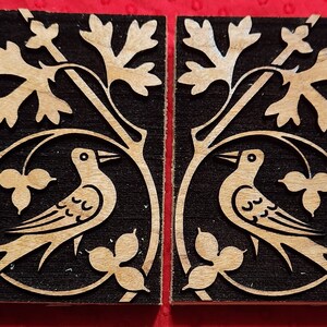 May include: Two wooden tiles with a black background and a brown design. Each tile features a stylized bird with a long tail and a leafy branch with a circular motif. The tiles are a decorative element for a home.