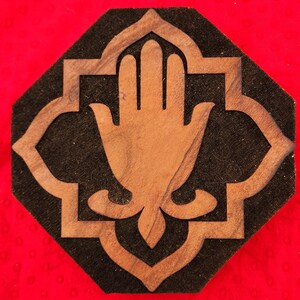 May include: A wooden Hamsa hand design, featuring a hand symbol within an ornate frame, is set against a black background. The design is placed on a red surface. The Hamsa hand is a symbol of protection and good fortune.