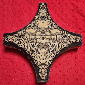 May include: A wooden, diamond-shaped decorative piece with an intricate design. The central motif features a stylized bird with outstretched wings, surrounded by floral and geometric patterns. The edges are black, and the background is a light wood color.
