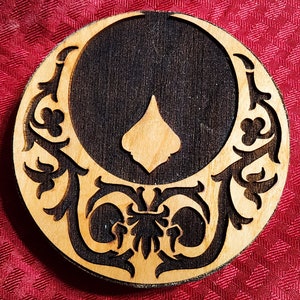 May include: A round wooden coaster with a dark brown center and intricate floral designs carved into the wood. The center of the coaster is a dark brown, almost black, and the floral designs are lighter brown.