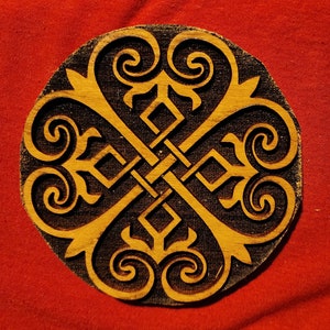 May include: A round wooden coaster with a black and gold intricate design. The design features a repeating pattern of swirls and knots.