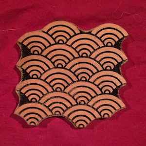 May include: A square wooden printing block featuring a repeating pattern of concentric semicircles in a wave-like arrangement. The design is deeply etched into the light-colored wood, with dark, recessed lines creating a high-contrast graphic effect.