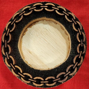 May include: A round wooden coaster with a black chain design around the edge. The coaster has a circular cutout in the center.