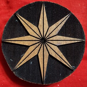 May include: A wooden coaster with a black background and a large, eight-pointed star design in a light brown color.