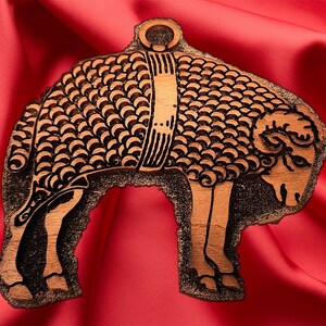 May include: A wooden cutout of a ram with a decorative design on its back. The ram is brown and black and has a ring at the top of its head.