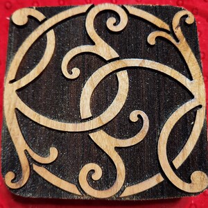 May include: A wooden block with a carved design of intricate swirls and loops. The design is in a light brown color and is set against a dark brown background.