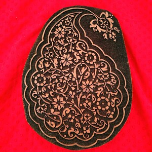 May include: A dark, paisley-shaped wooden block with intricate floral and vine patterns. The design is in a lighter brown color, set against a dark background. The block is on a bright red background.