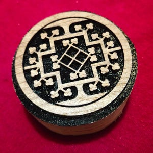 May include: A circular wooden stamp featuring a black recessed background and a light-colored natural wood surface. The design consists of a central diamond shape surrounded by a geometric pattern of branching lines with small clover-like ends.