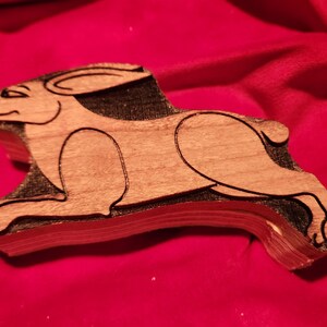 May include: A wooden cutout of a rabbit in a running pose. The rabbit is carved from light brown wood and has a dark brown outline. The cutout is on a red fabric background.