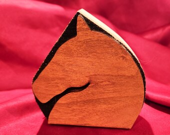 May include: A wooden horse head cutout with a dark brown outline. The horse head is carved from a light brown wood and is set against a red background.