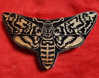 May include: A wooden moth-shaped carving with intricate details. The moth features a detailed design with black and brown tones. The wings and body are carved with precision, creating a striking contrast against the red background.