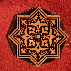 May include: A wooden star-shaped tile with a black and brown intricate design. The tile has a raised edge and is set against a red background.