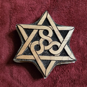 May include: A six-pointed star ornament crafted from light-toned wood, featuring an intricate, interlaced knotwork design at its center. The wooden star is layered over a textured black backing, creating a sharp contrast between the materials.