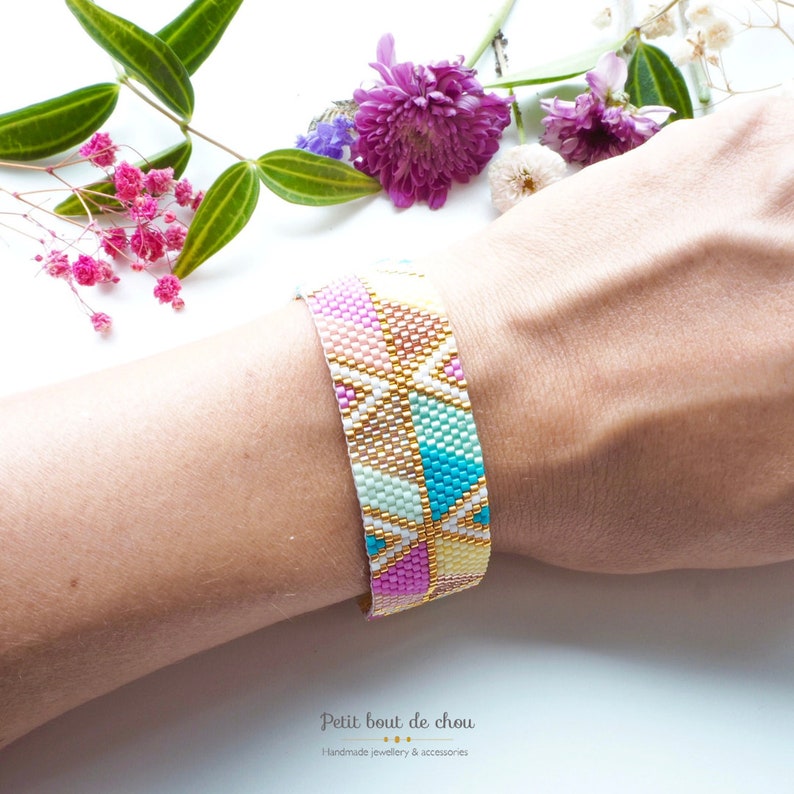 May include: A colorful beaded bracelet with a geometric pattern in shades of pink, blue, green, and gold. The bracelet is on a person's wrist.
