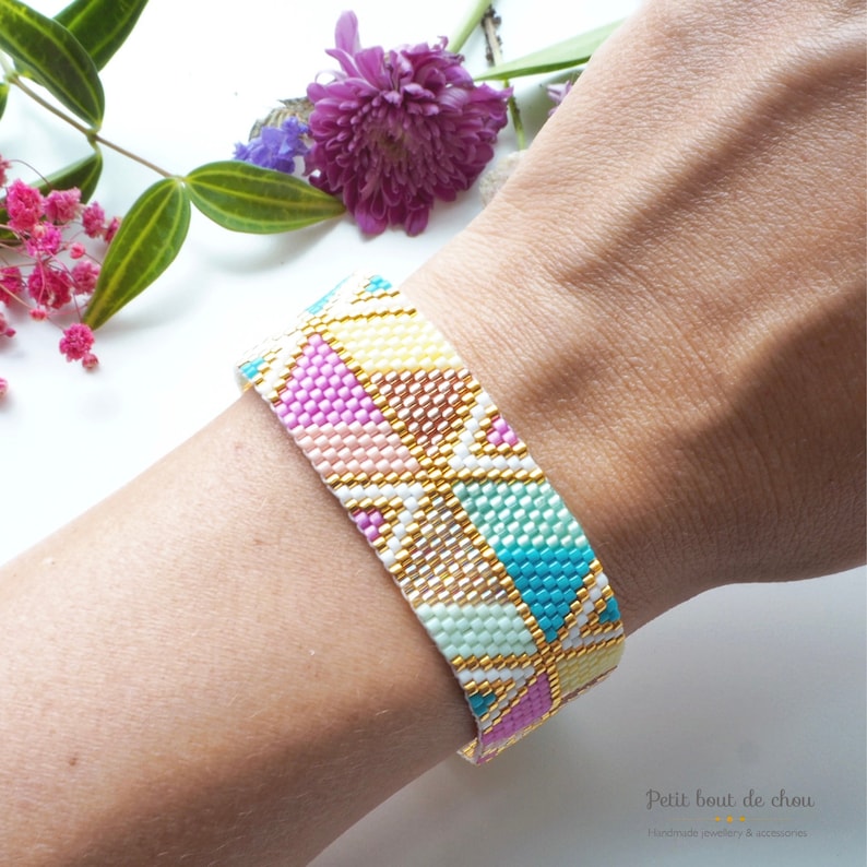 May include: A colorful beaded bracelet with a geometric pattern in shades of pink, yellow, blue, green, and white. The bracelet has gold accents and is worn on a person's wrist.