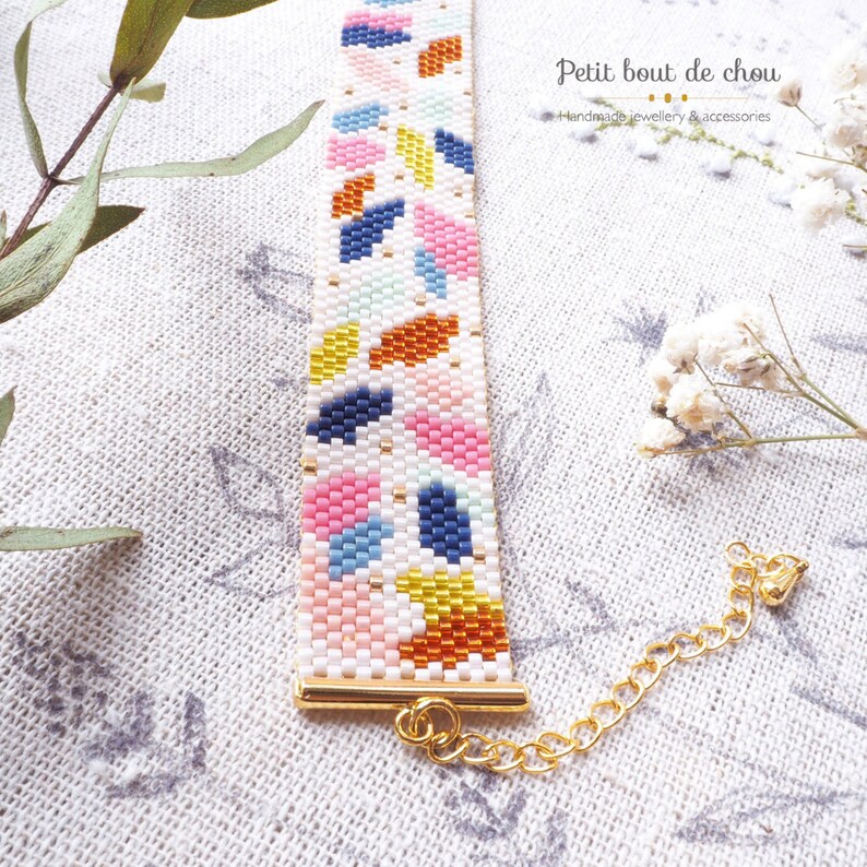 May include: A colorful beaded bracelet with a geometric pattern. The bracelet has a gold clasp and is made with white, blue, pink, yellow, and orange beads.