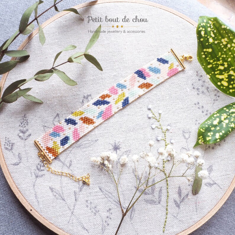 May include: A colorful beaded bracelet with a geometric pattern of pink, blue, yellow, and orange beads. The bracelet is made with a white background and has a gold clasp.