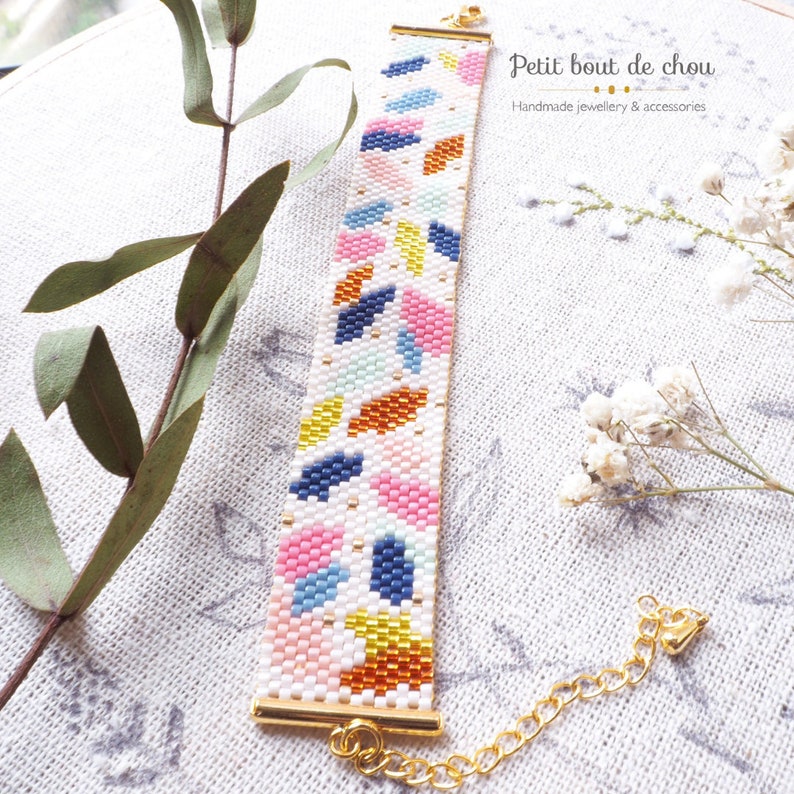May include: A colorful beaded bracelet with a geometric pattern in shades of pink, blue, yellow, and orange. The bracelet is made with small beads and has a gold clasp. The bracelet is on a white fabric background. "Petit bout de chou" is written in black text on a white background.