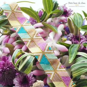 May include: A colorful beaded bracelet with a geometric pattern in shades of pink, yellow, blue, and green. The bracelet is made with small beads and has a gold thread running through it. The bracelet is photographed against a background of pink and purple flowers.