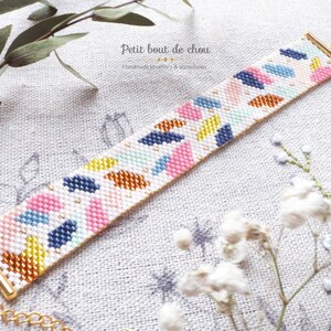 May include: A colorful beaded bracelet with a geometric pattern in shades of pink, blue, yellow, and orange. The bracelet is made with white beads and has a gold clasp.