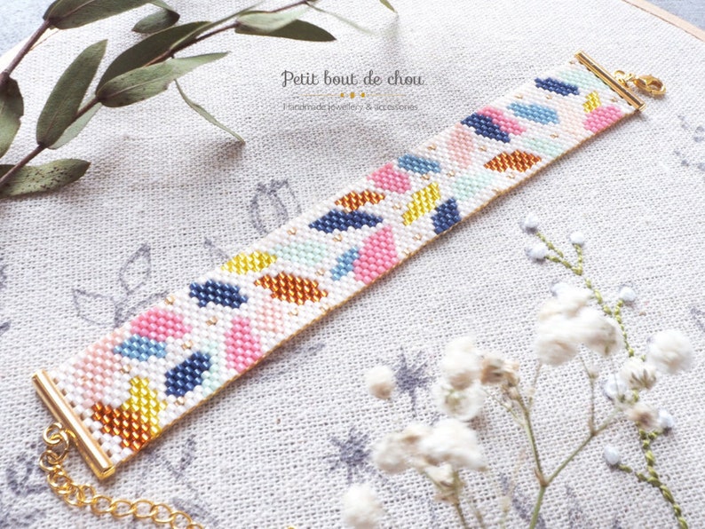 May include: A colorful beaded bracelet with a geometric pattern in shades of pink, blue, yellow, and orange. The bracelet is made with white beads and has a gold clasp.
