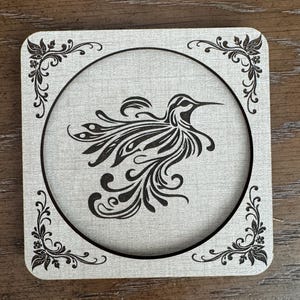 Könnte beinhalten: Quadratischer Holzuntersetzer mit einem Kolibri-Design. Der Untersetzer hat eine hellgraue Mitte mit einer schwarzen Kolibri-Illustration in einem schwarzen kreisförmigen Rahmen. Die Ecken haben dekorative schwarze Blumenmuster.