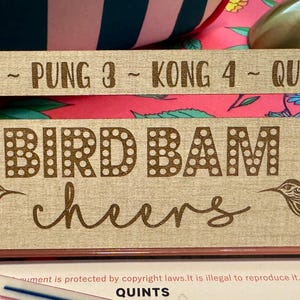 May include: A wooden Mahjong tile holder with the words "PAIR 2 - PUNG 3 - KONG 4 - QUINT 5" at the top. The words "BIRD BAM cheers" are in the center, with hummingbird designs on each side. The holder is beige with brown lettering.