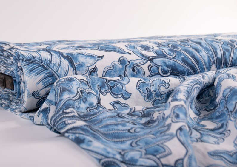 May include: A roll of fabric with a repeating blue and white pattern. The design features stylized cloud-like shapes and floral elements. The fabric is draped, showing the detailed print and texture.