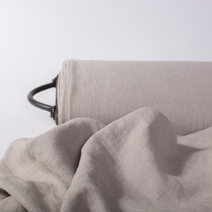 May include: A roll of light gray fabric with a black handle. The fabric is draped in the foreground, showing its texture. The material appears to be linen or a similar natural fiber, suitable for clothing or home decor.