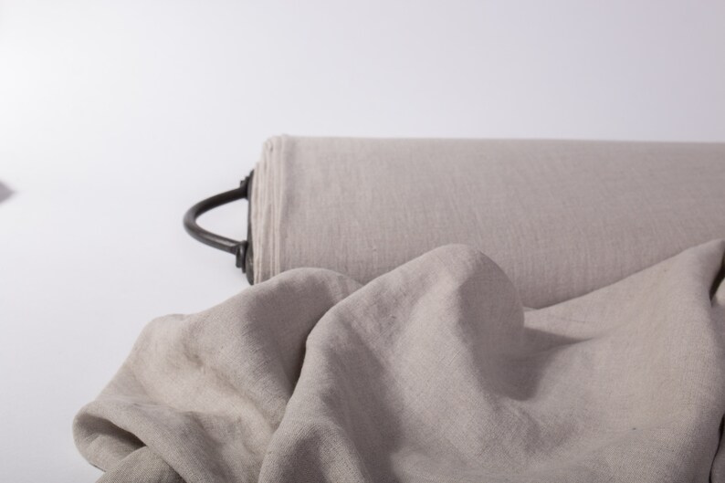 May include: A roll of light gray fabric with a black handle. The fabric is draped in the foreground, showing its texture. The material appears to be linen or a similar natural fiber, suitable for clothing or home decor.