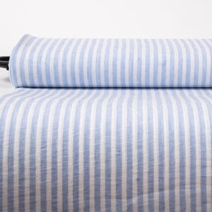 May include: A bolt of fabric featuring a repeating pattern of vertical light blue and white stripes. The textile is rolled onto a black cylindrical core, with a portion of the striped material draped downward to display the texture and color.